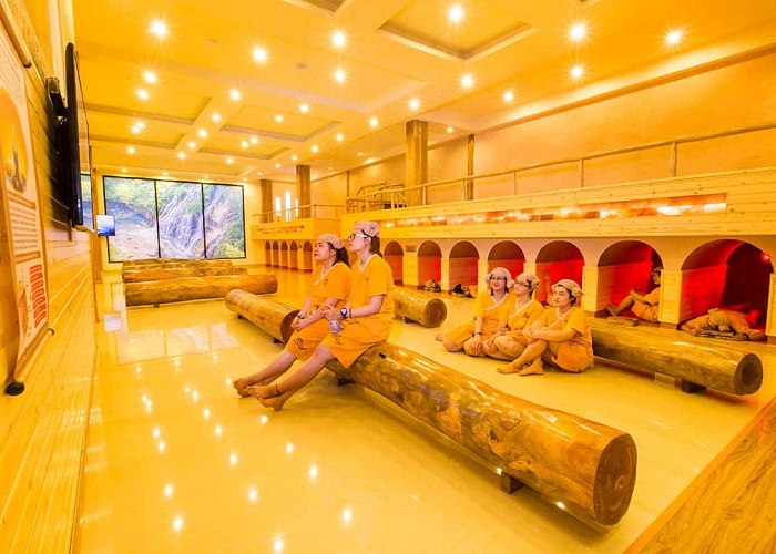 Sauna Korean with the person wearing the gold uniform, relaxing on wooden chairs, warm lighting.