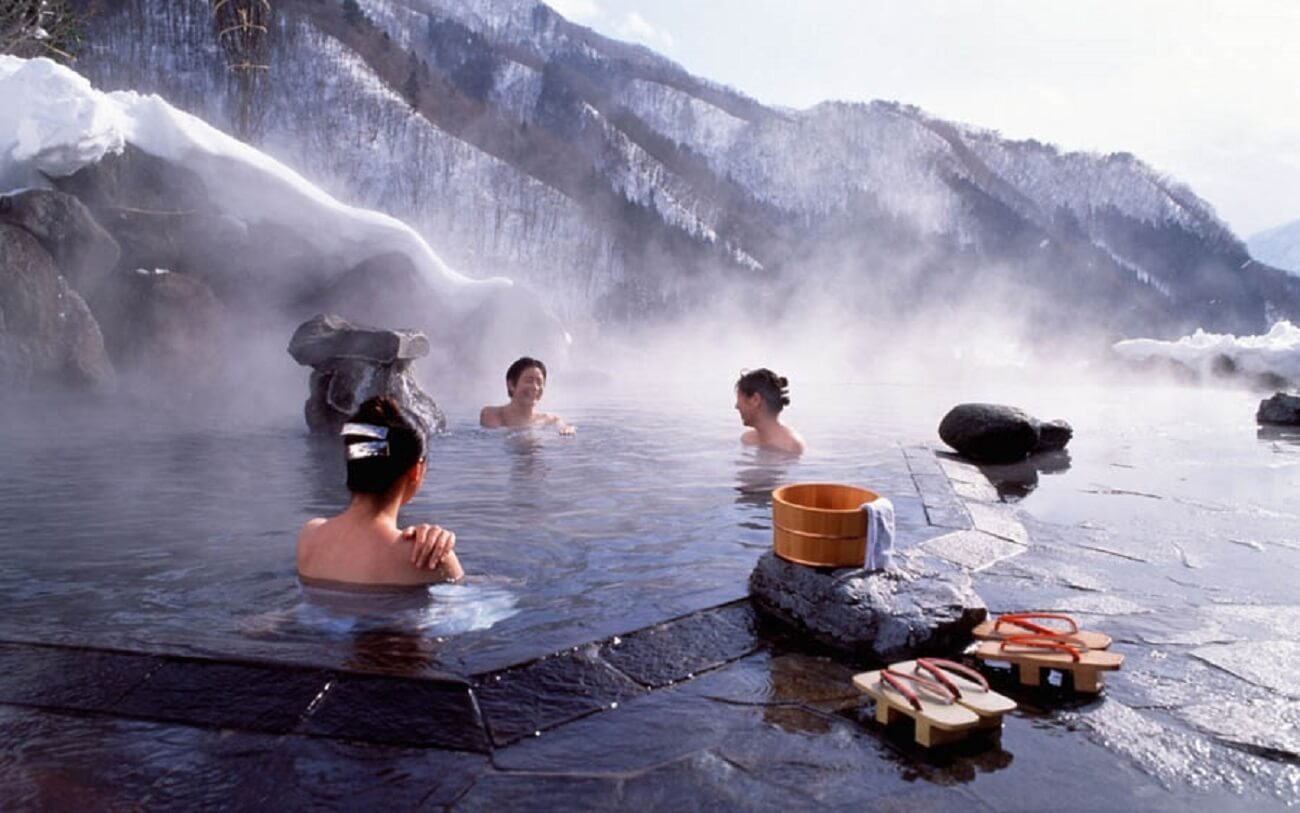 Người thư giãn trong onsen ngoài trời dưới trời tuyết ở Nhật Bản, hơi nước bốc lên tạo khung cảnh yên bình.