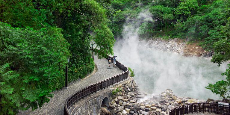 Đường mòn bên suối nước nóng giữa rừng xanh, hơi nước bốc lên tạo cảnh quan thiên nhiên thư giãn.