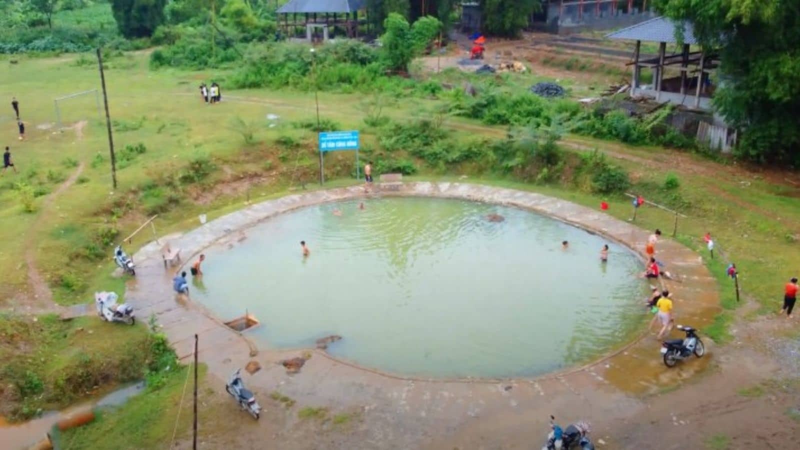 Hồ bơi ngoài trời nhỏ tại nông thôn Việt Nam với một số người đang bơi, xung quanh là cây xanh và xe máy.