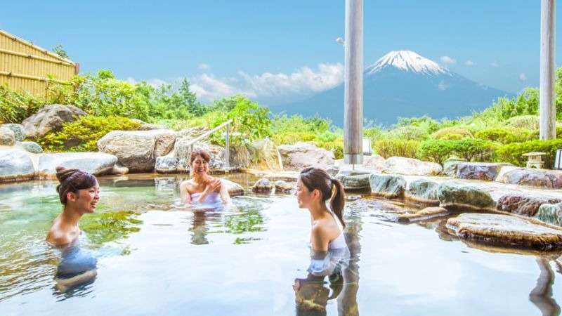 Ba người phụ nữ tận hưởng tắm onsen ngoài trời với cảnh núi Phú Sĩ tuyệt đẹp phía xa, tạo nên khung cảnh thiên nhiên thư giãn.