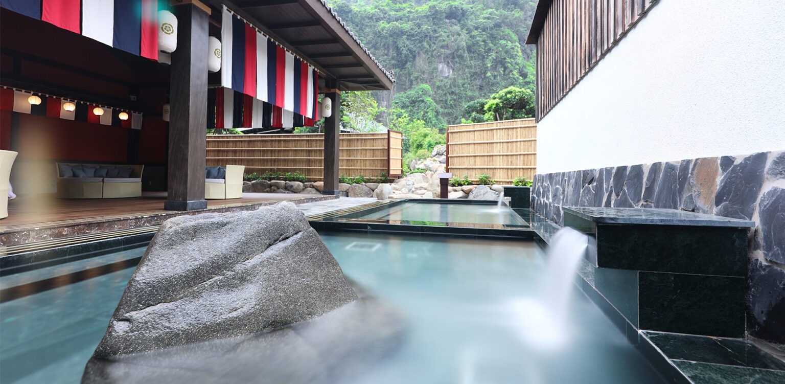 Khu vườn Nhật Bản với onsen, cây cối xanh mát và nhà gỗ truyền thống, tạo không gian thư giãn thanh bình.