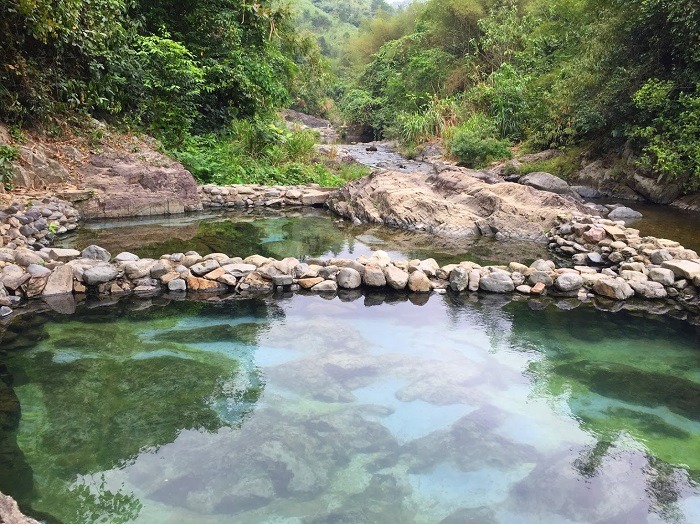 Suối khoáng nóng với nước trong xanh, đá xếp tự nhiên và cây xanh xung quanh, tạo cảm giác thư giãn và yên bình.