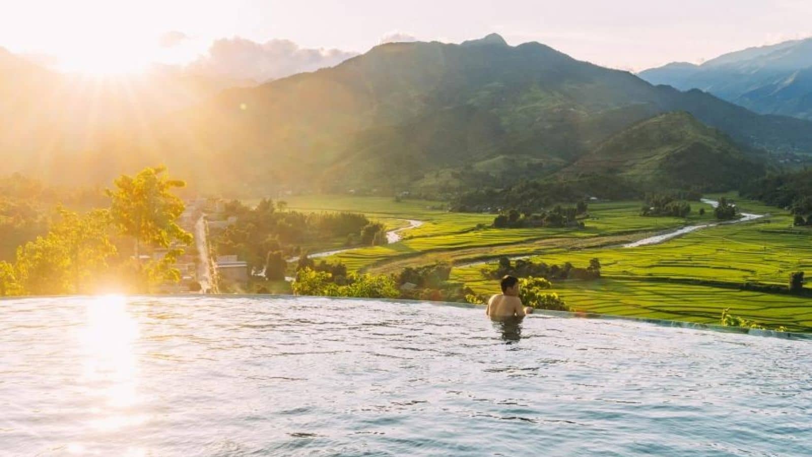 Người trong bể bơi vô cực nhìn ngắm cánh đồng và núi đồi dưới ánh hoàng hôn, tạo khung cảnh yên bình.