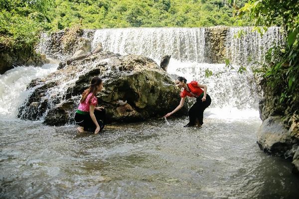 Hai người phụ nữ mặc trang phục truyền thống chơi đùa dưới thác nước nhỏ ở vùng núi xanh mát.