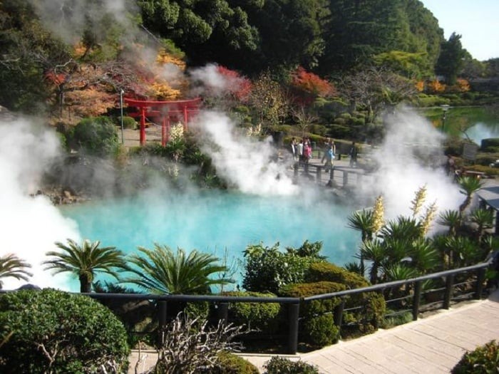 Hồ nước nóng tự nhiên với làn khói bốc lên, cây xanh và cổng Torii đỏ trong khung cảnh mùa thu yên bình.