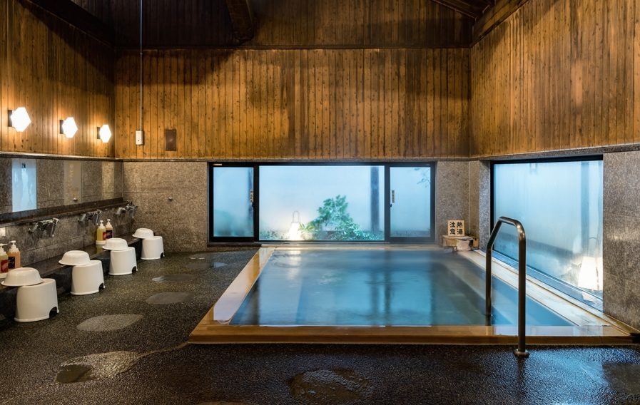 Phòng tắm onsen Nhật Bản sang trọng với bồn nước nóng, tường gỗ truyền thống và cửa sổ lớn, mang lại không gian thư giãn.