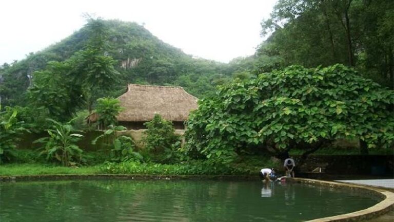 Cảnh núi đồi và nhà tranh bên hồ nước xanh mát, với cây cối tươi tốt bao quanh, tạo nên khung cảnh yên bình.