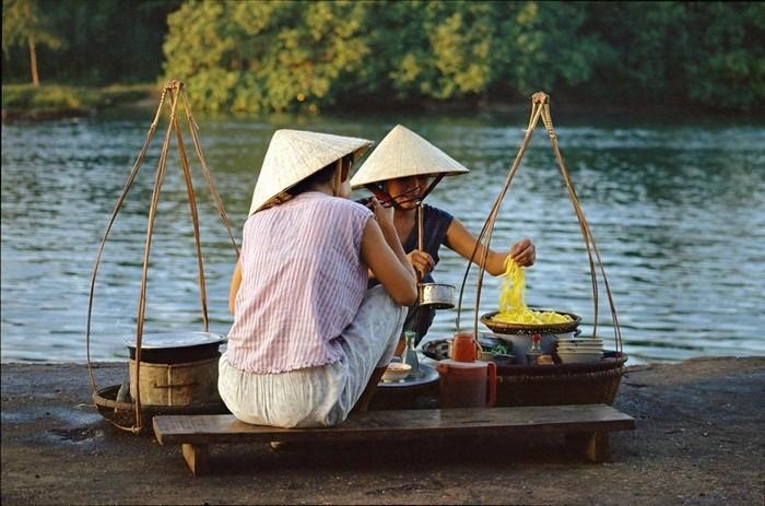 Người phụ nữ đội nón lá bán hàng bên sông với gian bếp nổi, tái hiện hình ảnh đời sống nông thôn Việt Nam.