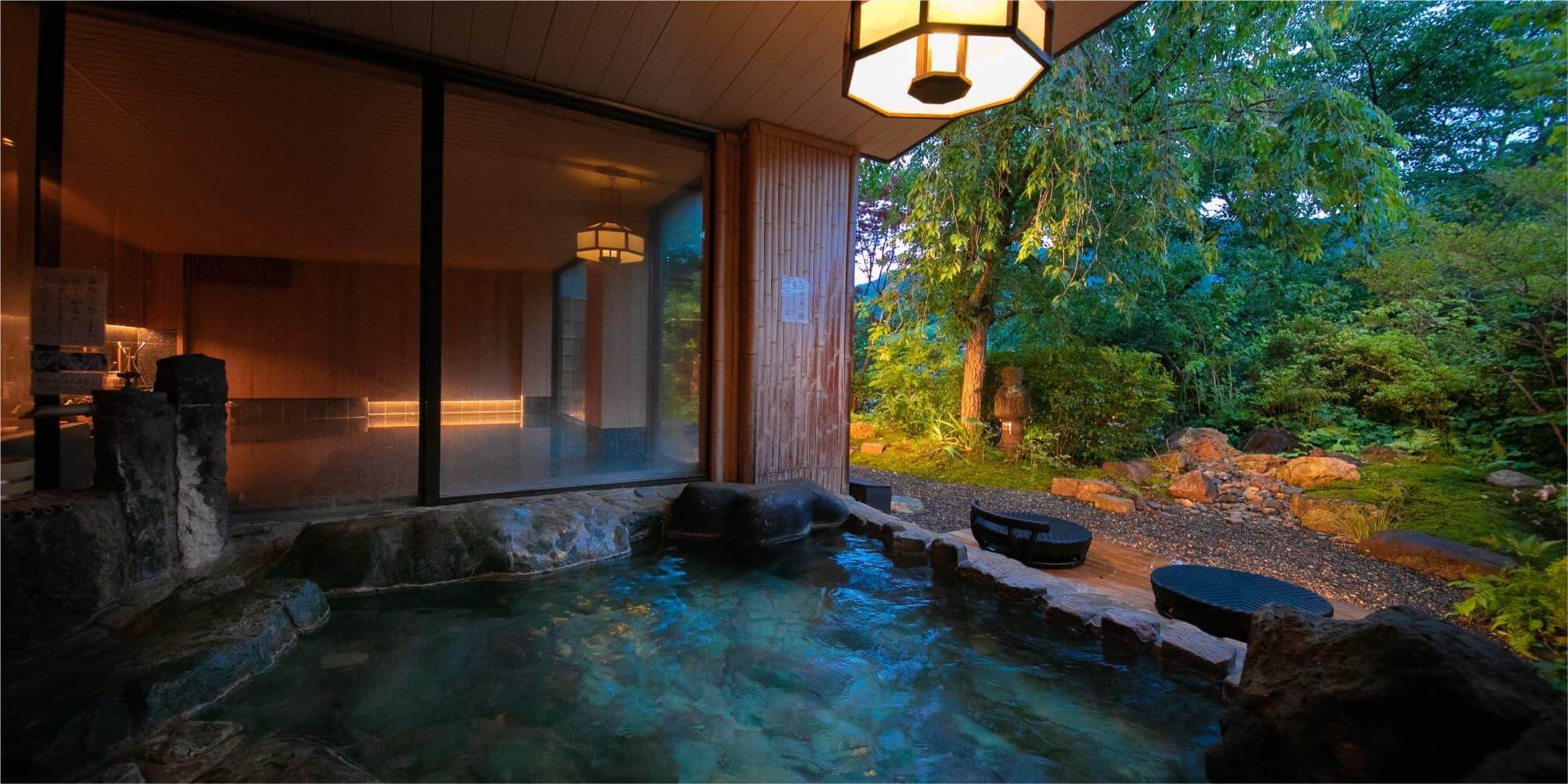 Phòng tắm onsen nhìn ra khu vườn xanh mát vào buổi tối với đèn chiếu sáng ấm áp mang lại không gian thư giãn.