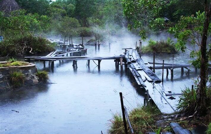 Cầu gỗ cũ bên hồ nước khói mờ, bao quanh bởi cây cối xanh tốt, tạo cảm giác huyền bí và yên bình.